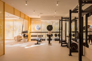 A man performing overhead barbell press in a bright, modern home gym featuring golden perforated metal walls, black power racks, dumbbell storage, and large mirrors.