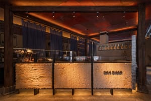Modern Japanese-inspired cafe interior featuring a massive textured stone counter with backlit 'SAN SHAN' signage, dark timber structural beams, and warm ambient lighting.