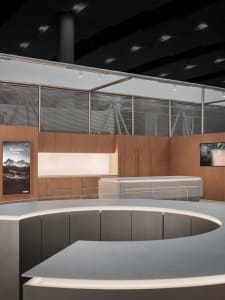 Modern luxury boutique interior featuring a large curved grey reception island in the foreground, warm oak cabinetry and backlit display wall in the background, with large windows covered by horizontal blinds.