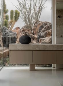 Modern minimalist bathroom featuring a floating travertine vanity with a black ceramic vessel, large glass window overlooking dramatic desert rocks and cacti.