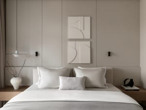 Modern minimalist bedroom with neutral beige walls, upholstered bed with white and grey bedding, two abstract white wall sculptures, wooden nightstands, and a hanging pendant lamp.