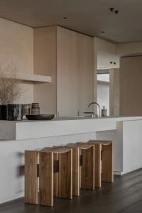 Modern minimalist kitchen interior with raw concrete island, curved walnut wooden stools, matte beige cabinetry, and dried floral arrangement.