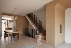 Modern dining room featuring a solid oak table and chairs, three pendant lights, light oak wall paneling, a black metal railing staircase, and a curly-haired dog standing on light wood flooring.