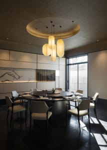 Modern Chinese-style dining room with a large round black marble table, eight cream upholstered chairs, textured wall panels, ink wash painting, and a sculptural cluster of cylindrical pendant lights hanging from a circular ceiling feature.
