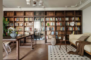 Modern living room featuring a full-wall walnut wood bookshelf, wooden desk, cream sofa, and black-and-white geometric rug under natural light.