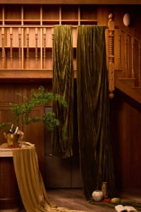 Interior view of a luxurious wooden staircase with turned balusters, featuring floor-to-ceiling olive green velvet curtains, warm walnut paneling, and a styled round table with champagne bucket and greenery in the foreground.