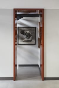 Modern minimalist hallway featuring a large framed black and white nude photograph, rich walnut wood door frames, and dramatic diagonal light patterns on white walls.