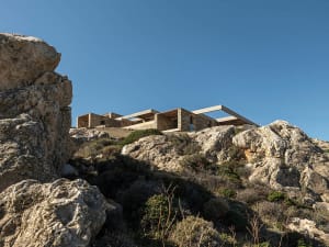 Modern stone and concrete house built into a rocky cliffside with dramatic overhangs and open terraces, surrounded by Mediterranean scrub vegetation under a clear blue sky.