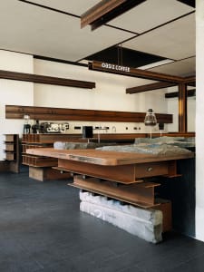 Modern cafe interior featuring a large coffee counter made from weathered corten steel beams and thick stone slabs, with matching industrial shelving and dark tiled flooring.