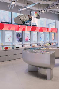 Modern cosmetics store interior featuring a large curved grey display island, red accent wall with circular patterns, product testing stations, and large windows with city views.