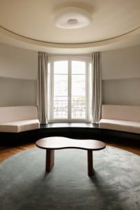Modern minimalist living room featuring a curved bay window with built-in upholstered benches, a free-form walnut coffee table, and a large round blue-gray rug under a circular ceiling light.