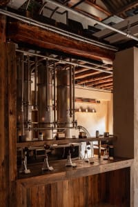Interior view of an industrial-style distillery bar featuring large copper distillation columns, heavy reclaimed wood beams and counters, exposed metal pipes, and laboratory glassware under warm lighting.