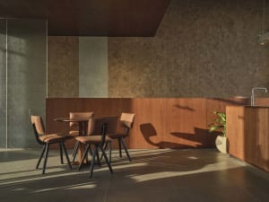 Modern minimalist dining area featuring a round walnut table, four terracotta upholstered chairs with black metal frames, wood-paneled walls, and dramatic natural light creating long shadows on a dark tiled floor.