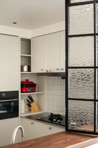 Modern minimalist white kitchen featuring handleless matte cabinets, black induction cooktop, open shelving with red multi-cooker and knife block, and a textured glass partition on the right.