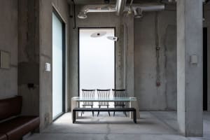 Industrial-style dining area featuring raw concrete walls, a glass dining table with black spindle-back chairs, a black bench, and two modern pendant lights suspended from an exposed ceiling with visible ductwork.