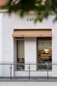Street view of a modern white cafe named CALI with beige awnings, large glass windows showing customers inside, and a metal railing in the foreground.