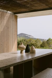 Speckled terrazzo counter with three rustic ceramic vases in beige, black and light terracotta, set against a wooden slat wall and panoramic mountain view under a thatched roof.