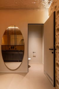 Modern minimalist bathroom with warm terracotta plaster walls, a large oval mirror reflecting a dark wood vanity, and a sculptural curved wall with circular cutouts.