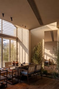 A tranquil dining area featuring a dark wood table, woven rattan and fabric dining chairs, colorful wooden side tables, and tall bamboo plants, with strong sunlight casting striped shadows on beige plaster walls.