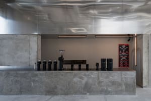 Modern minimalist coffee bar featuring a massive concrete counter, stainless steel backsplash with wavy texture, black coffee equipment, and a digital menu board under linear lighting.