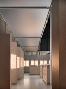 Long perspective view down a minimalist jewelry boutique interior featuring floor-to-ceiling oak veneer cabinets with integrated backlit display cases showcasing jewelry illustrations and products.