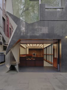 Modern bakery storefront featuring a dramatic angular facade made of weathered corten steel and concrete, with an open view into a wood-lined interior displaying fresh pastries.