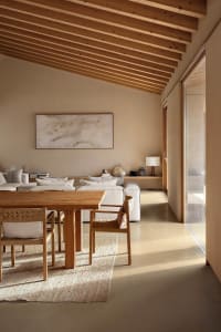 Modern minimalist dining area with solid oak table and woven rattan chairs on a textured rug, white sectional sofa in the background, exposed wooden beam ceiling and large abstract painting on the wall.