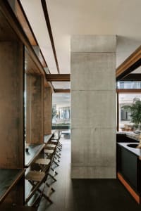 Narrow industrial cafe interior featuring a massive raw concrete pillar, long wooden counter with folding wooden stools, dark metal accents and natural light from large windows.
