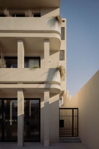 Exterior view of a modern multi-story concrete building featuring rounded balconies, vertical pillars, large glass doors, and potted grasses under a clear blue sky.