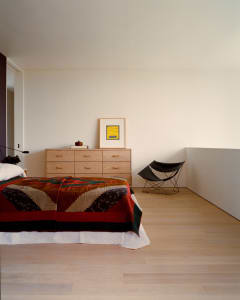 Minimalist bedroom interior with a bed covered in a colorful geometric quilt, light oak dresser against a white wall, framed yellow artwork, and a black butterfly chair in the corner.