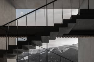 Modern concrete staircase with black metal railings overlooking snow-capped mountains and forested valleys.