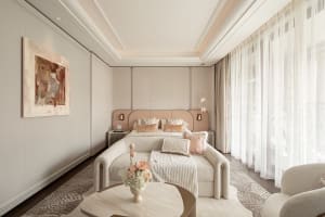 Modern luxury bedroom with blush pink upholstered bed, cream boucle sofa, marble coffee table, abstract wall art, and sheer white curtains allowing soft daylight.