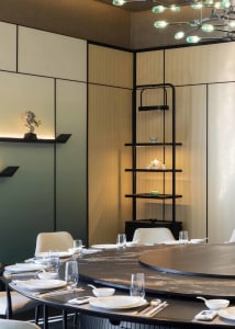 Modern Chinese-fusion dining room with a large round black marble table, lazy Susan, white upholstered chairs, perforated beige wall panels, and a sculptural green glass chandelier.