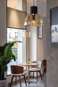 Modern minimalist cafe interior featuring a round wooden table, walnut and upholstered dining chairs, large potted plant, and warm pendant lights by a window with blinds.