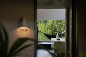 View through a doorway into a minimalist concrete reading nook featuring a large window overlooking a dense green forest with a black hammock suspended outside.