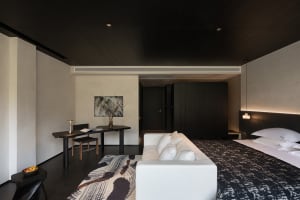 Modern minimalist bedroom featuring dark wood ceiling and walls, a large bed with white linens, a white sofa, an abstract black and white rug, and a built-in desk area with artwork.