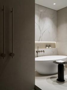 Modern minimalist bathroom featuring a white freestanding oval bathtub, large marble tile walls, recessed lighting, and a black cylindrical side table with folded white towels.