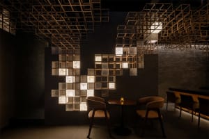 Modern restaurant interior featuring a large illuminated grid wall made of wooden cubes and textured glass panels with dried flowers, paired with round wooden tables and tan leather chairs under a dramatic geometric ceiling installation.