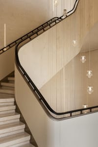 Modern curved travertine staircase with black metal handrail and multiple crystal pendant lights hanging in a vertical cascade against a textured beige wall.