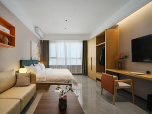 Modern hotel bedroom with teal velvet headboard, oak wardrobe and desk, beige sofa, and large window with sheer curtains.