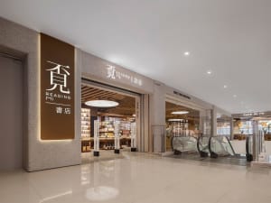 Modern bookstore entrance featuring a large illuminated brown sign with Chinese characters for Reading Mi, textured terrazzo walls, polished reflective floor, and escalators visible in the background of a spacious shopping mall.