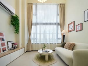 Modern minimalist living room with a sage green boucle sofa, curved white coffee table on a green rug, large window with sheer curtains, and built-in low cabinet displaying art and plants.