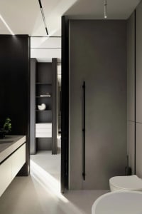 Modern minimalist bathroom interior with dark wood cabinetry, white floating vanity, built-in open shelving, matte black door, and white ceramic toilet under dramatic natural light.