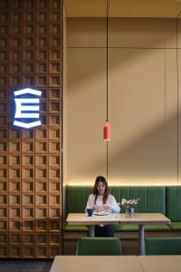 Woman dining alone at a light wood table in a modern restaurant booth with green upholstery, a coral pendant lamp, and a striking textured wooden wall featuring a glowing neon logo.