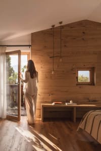 Woman in cream outfit standing by open glass door in a modern wooden bedroom with oak walls, pendant lights, and mountain view.