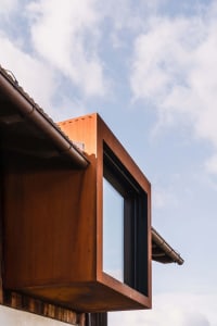 Low-angle view of a rust-colored corten steel cantilevered window box extending from a building roofline with blue sky and clouds in the background.