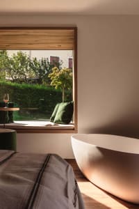 Modern bathroom interior featuring a white freestanding bathtub, built-in oak window seat with green cushions, wooden flooring, and a large window overlooking a lush garden.