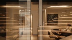 Modern dining room viewed through multiple layers of curved glass partitions with warm ambient lighting, abstract black artwork, and a white sculpture in the background.