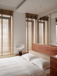 Modern minimalist bedroom with a large red travertine stone headboard, white bedding, wooden Venetian blinds, and a sculptural white table lamp.