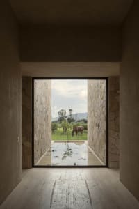 View from inside a minimalist concrete and stone room looking out through a large opening to a shallow reflecting pool, stone walls, green pasture, and a brown horse grazing with distant mountains.